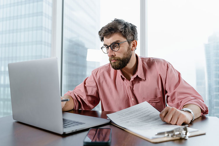 Man with glasses and pink dress shirt at desk with a laptop and income tax documentation on a clipboard