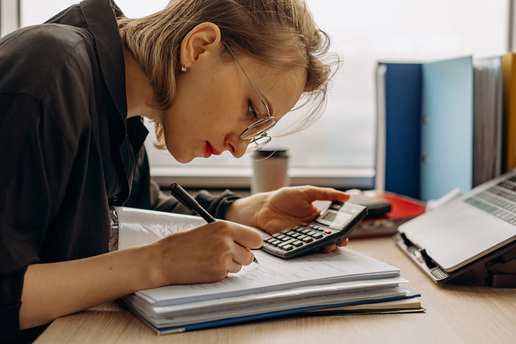 Picture of woman in glasses holding calculator and writing down numbers for accounting
