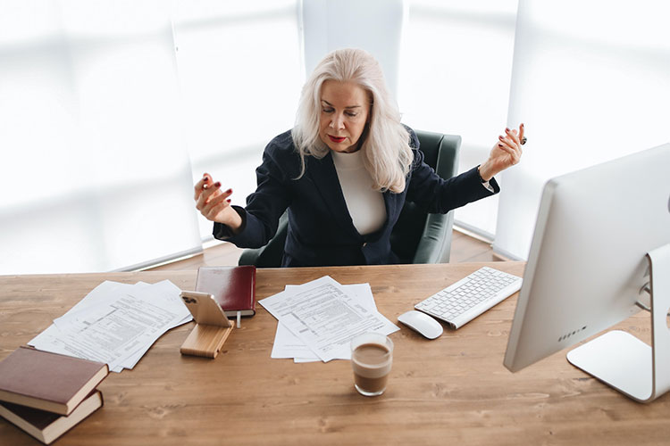 Woman with gray hair in a business suit sitting at work desk with coffee, computer and tax documents