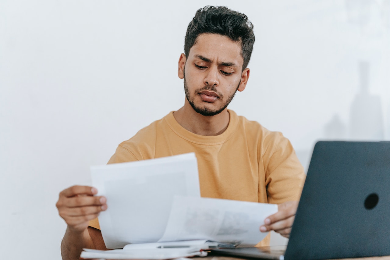 Picture of young man in yellow sweater reviewing tax papers in front of laptop