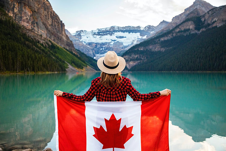 woman in plaid shirt and hat, holding a Canadian flag looking out at lake and mountains