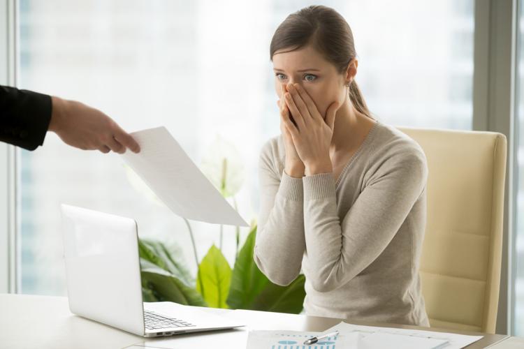 Distraught woman at her desk covering her mouth with her hands being handed a paper from someone wearing a suit