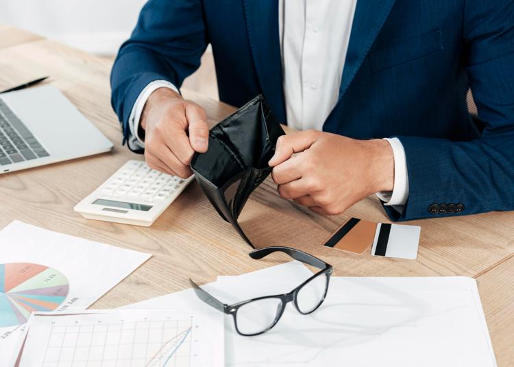 Closeup of a man in a suit at a desk with papers and a laptop opening an empty wallet