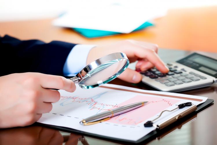 Closeup of a person wearing a suit holding a magnifying glass to a chart with one hand while using a calculator with the other.