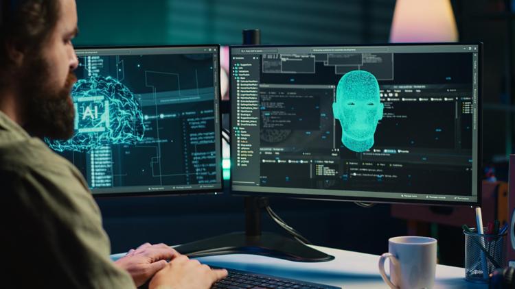 Man working at a desktop computer with a digital human and brain appearing on the double monitors.