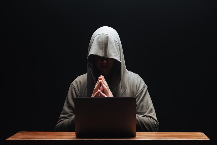 A no-face, hooded hacker in shadow in front of a laptop with his fingers tented.