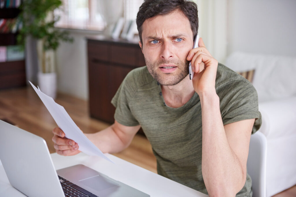 Confused man on the phone and holding papers in front of a laptop in a desk
