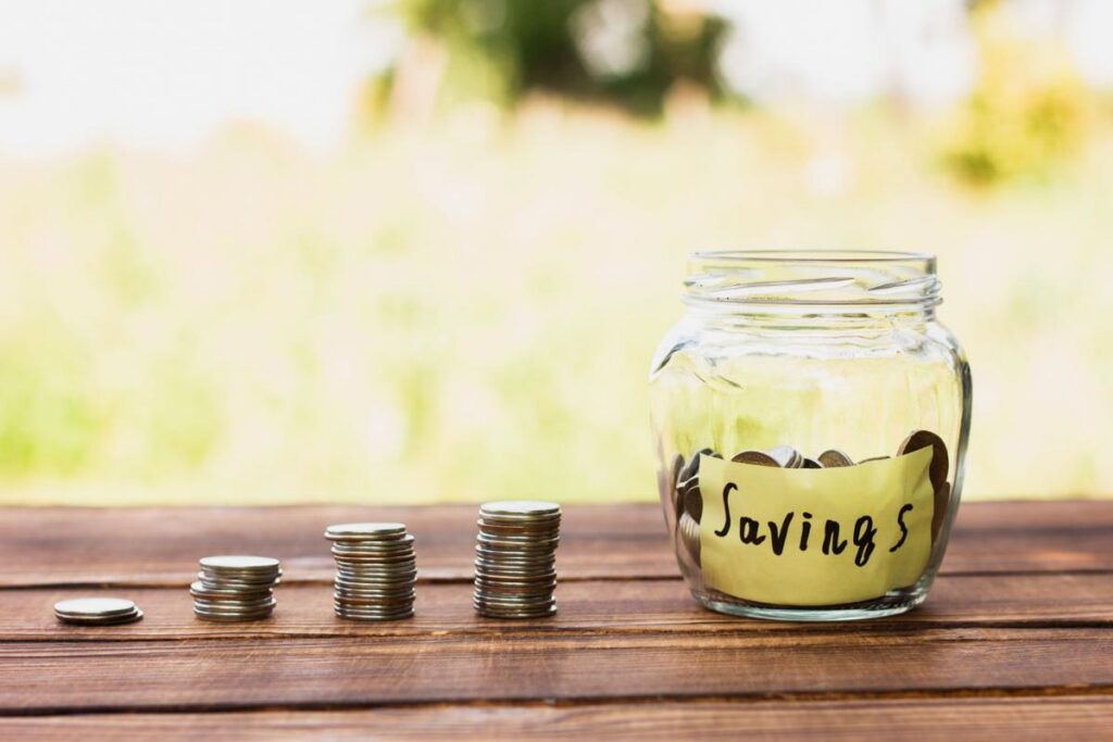 A Jar labelled Savings on a table beside four variously-sized stacks of coins.